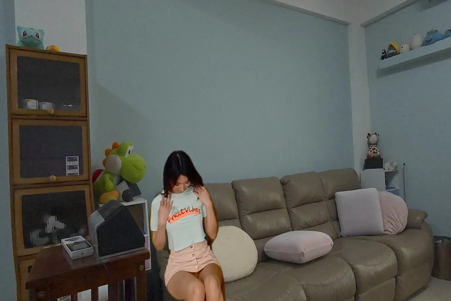 アジアの女の子が薄緑のTシャツとピンクのミニスカートを着て、ソファに座りシャツを引っ張りながらうつむいている。背景にはぬいぐるみやクッションがある
